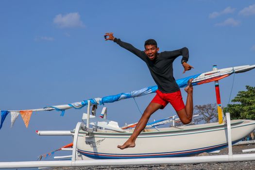 Happy young man jumping on the beach. Young cheerful Indonesian jumps. A teenager in a good mood jumps in the air. In the background a fishing boat. Sunny tropical day with blue sky.