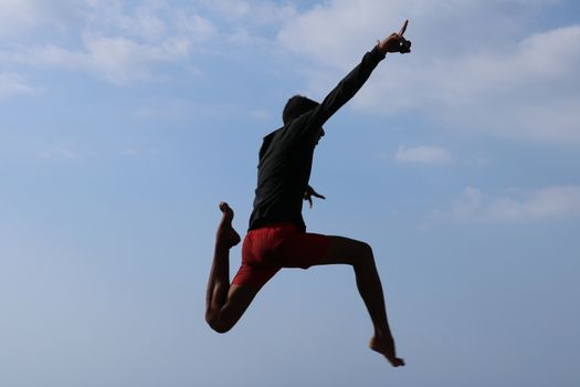 Happy han jumping above blue sky background. Young cheerful Indonesian jumps. A teenager in a good mood jumps in the air. Sunny tropical day.
