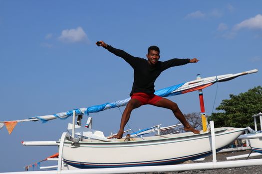 Happy young Indonesian man jumps on the beach. A teenager in a good mood jumps in the air. Sunny tropical day with blue sky.