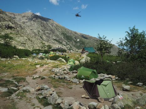 France, Corsica, Corsician Alps, June 19, 2017: helicopter dropping off supplies for mountain camp Refuge de Pietra Piana on famous hiking trail GR20, rocks, tents, tree and blue sky background