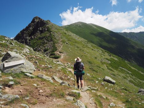 hiker man in hat with heavy backpack walking on GR 20 famous trail in corsician alpes, scenery of high mountain green meadow with blue sky background