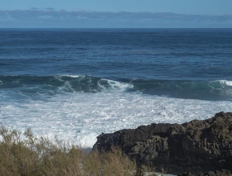 Volcanic rock formation, cliffs of black lava on the rocky shore with crushing white waves over the Atlantic Ocean. Blue sky background. La Palma, Canary Islands