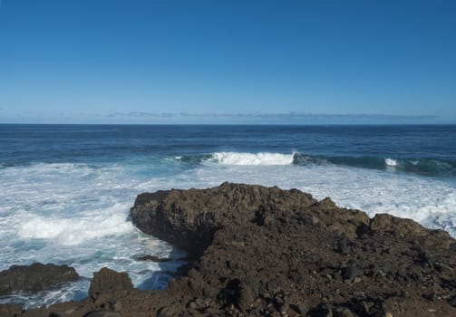 Volcanic rock formation, cliffs of black lava on the rocky shore with crushing white waves over the Atlantic Ocean. Blue sky background. La Palma, Canary Islands