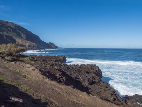 Volcanic rock cliffs of black lava on the rocky shore with crushing white waves over the Atlantic Ocean. Blue sky background. La Palma, Canary Islands