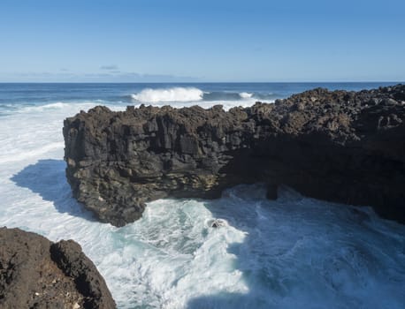 Volcanic rock formation, cliffs of black lava on the rocky shore with crushing white waves over the Atlantic Ocean. Blue sky background. La Palma, Canary Islands