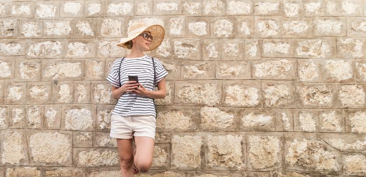 Beautiful young female tourist woman standing in front of old textured stone wall at old Mediterranean town, smiling, holding, smart phone to network on vacationes. Copy space.