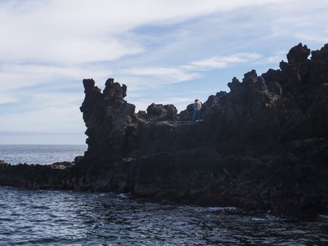 Back side of older man, local fisherman in hat standing on the edge of lava rock cliff with fishing rod at coast of atlantic ocean at La Palma island, Spain.