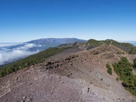 Volcanic landscape with lush green pine trees, colorful volcanoes and lava rock field along path Ruta de los Volcanes, hiking trail at La Palma island, Canary Islands, Spain, Blue sky background.