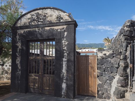 Old lava stone and wooden rustic door, blue house, white apartments, palm trees, green hills and blue sky at Canary islands village