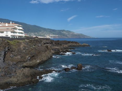 View on sea shore with lava rock cliffs at village Los Cancajos, ocean and green hills. Blue sky background. La Palma, Canary Islands, Spain.