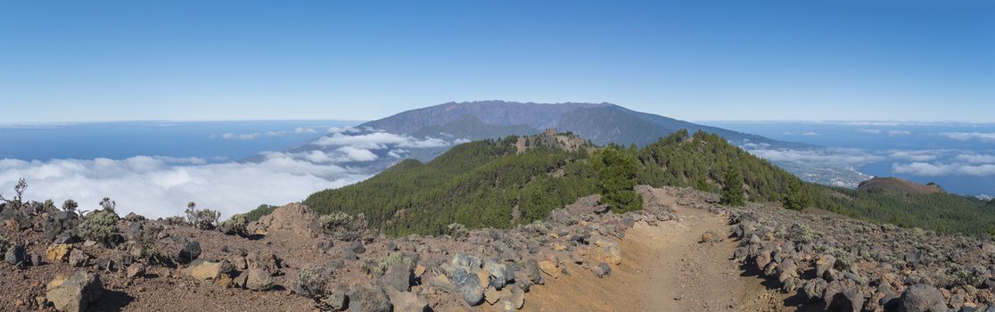 Panoramatic view of volcanic landscape with lush green pine trees, colorful volcanoes and white clouds at path Ruta de los Volcanes, hiking trail. La Palma, Canary Islands, Spain, Blue sky background.