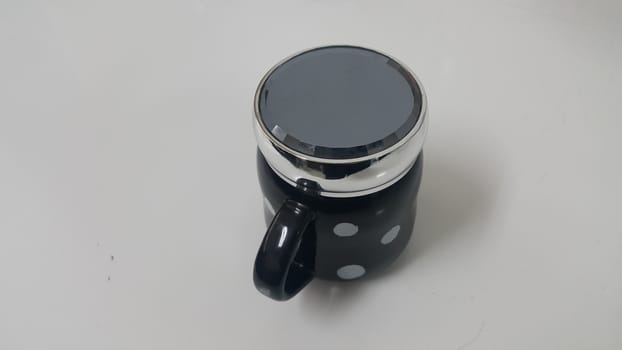 Beautiful coffee black mug with white dots placed over white floor with black lid on it. Closeup perspective view of a mug on white desk.