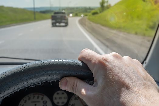 the driver hand on the steering wheel of a car that is passing on a country highway