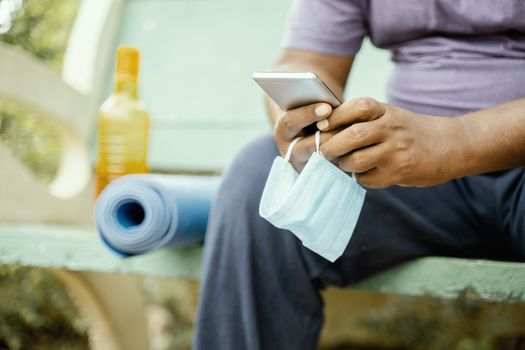 Man using mobile with mask in hand at park during morning yoga or workout with mat - concept of coronavirus or covid-19 advice, protection or to prevent from spreading by wearing mask at outdoor