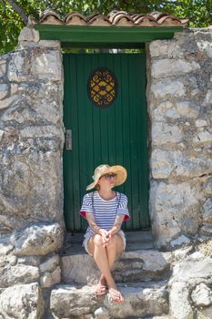 Beautiful young female tourist woman sitting and resting on vinatage wooden doorstep and textured stone wall at old Mediterranean town, smiling, holding, using smart phone to network on vacationes.