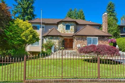 Luxury residential house with metal fence in front in suburb of Vancouver. Beautiful house with green lawn in front on blue sky background