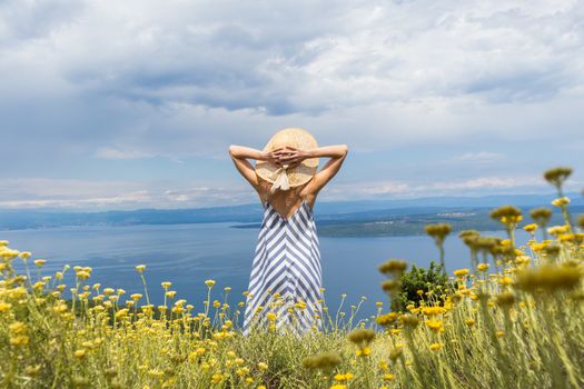 Rear view of young woman wearing striped summer dress and straw hat standing in super bloom of wildflowers, relaxing while enjoing beautiful view of Adriatic sea nature, Croatia.