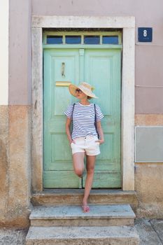 Beautiful young female tourist woman wearing sun hat, standing and relaxing in shade in front of turquoise vinatage wooden door in old Mediterranean town while sightseeing on hot summer day.