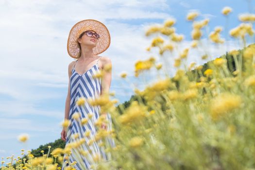 Young woman wearing striped summer dress and straw hat standing in super bloom of wildflowers, relaxing while enjoing beautiful nature of of Adriatic sea coastal nature of Croatia.