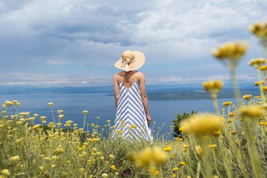 Rear view of young woman wearing striped summer dress and straw hat standing in super bloom of wildflowers, relaxing while enjoing beautiful view of Adriatic sea nature, Croatia.