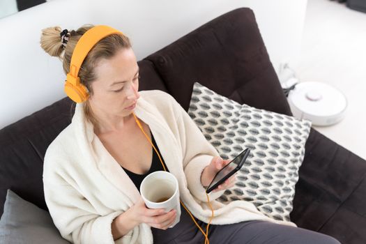 Woman at home relaxing on sofa couch drinking tea from white cup, listening to relaxing music while reading emails and using social media on mobile phone device. Stay at home. Social distancing.