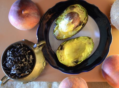 two avocado halves on a plate, three ripe peaches and a small copper jug of whole leaf tea and a coconut