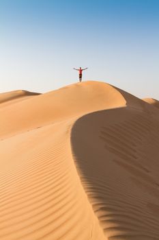 Male traveler standing on the top of dune, arms up to the sky, while traveling sand dessert in Oman. Travel adventure concept.