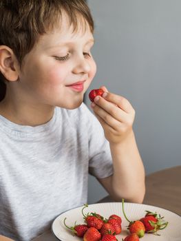 Four-year-old boy eats fresh strawberry with relish. Happy smiling child eats organic strawberry at the kitchen table. Vertical
