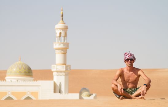 Man traveling in Oman, topless wearing a turban and shorts, sitting on send dune in fron of mosque in desert Oman.