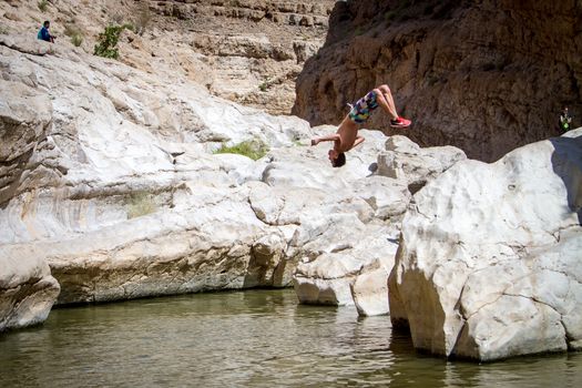 Sporty guy doing backflip off cliff into lake in an oasis in middle of Oman desert.