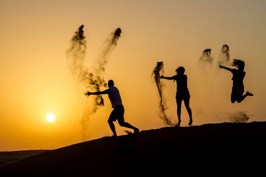 Silhouette of happy traveling people jumping on sand dune and throwing sand in the air in golden sunset hour.