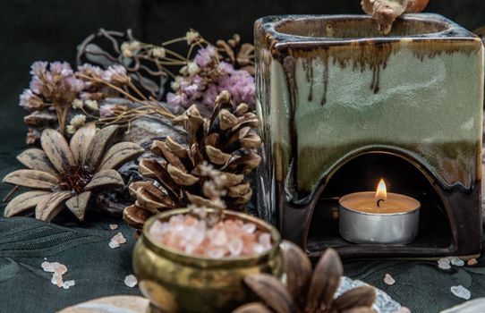 Beauty composition with Burning candle aroma oil lamp and Dry flower petals and Pine cones, Natural pine wood disc, Stones and Shells, Himalayan salt on dark background. Aromatherapy concept.