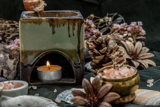 Beauty composition with Burning candle aroma oil lamp and Dry flower petals and Pine cones, Natural pine wood disc, Stones and Shells, Himalayan salt on dark background. Aromatherapy concept.