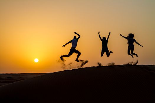 Silhouette of happy traveling people jumping on sand dune and throwing sand in the air in golden sunset hour.