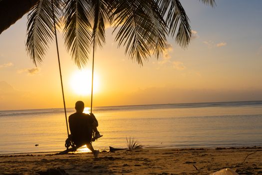 The guy enjoys the sunset riding on a swing on the ptropical beach. Silhouettes of a guy on a swing hanging on a palm tree, watching the sunset in the water