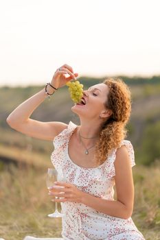 Beautiful young red-haired tender girl in a white sundress, eats grapes and drinks wine on the nature