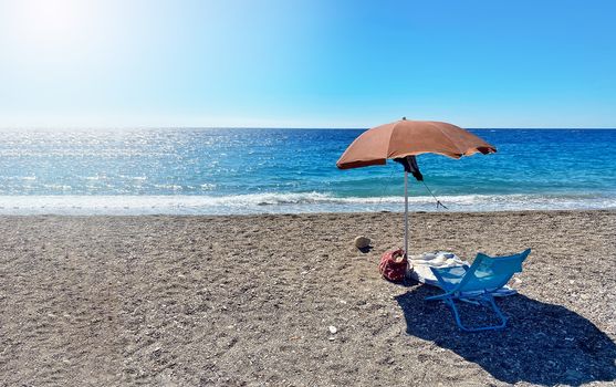 A deckchair under an umbrella facing the blue sea in a stony beach in Sicily, Italy. Summer holidays in Italian seaside resorts. Relax