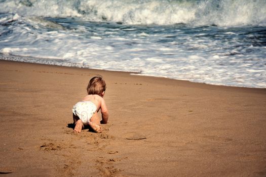 Infant boy crawling on sand towards ocean wave. Baby child crawl on all fours on tropical beach reaching waterfront rear view. Toddler curiosity of seaside at summertime. Infancy exploration. 