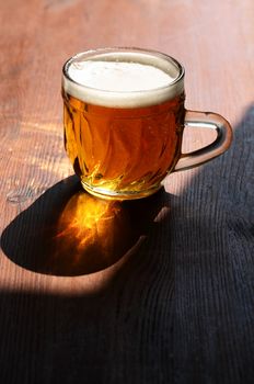 Mug of beer with foam on wooden table under sunlight