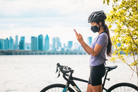 Biking cyclist using mobile phone for contact tracing app wearing COVID-19 face mask as coronavirus prevention while riding road bike outside on city travel vacation eco-tourism.