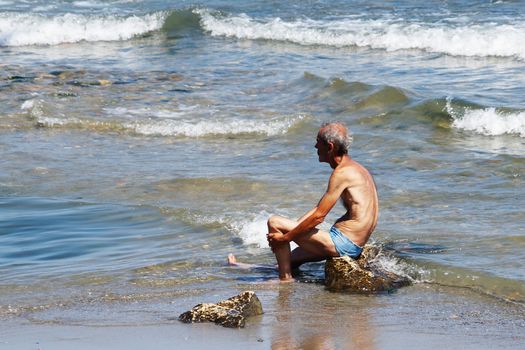 Varna, Bulgaria - July, 19, 2020: Elderly thin male yogi sits on a rock in the sea and contemplates