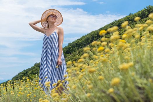 Young woman wearing striped summer dress and straw hat standing in super bloom of wildflowers, relaxing while enjoing beautiful nature of of Adriatic sea coastal nature of Croatia.