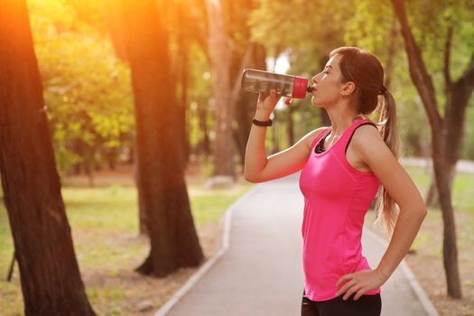 Beautiful fitness athlete runner woman drinking water in the park. Portrait face of a young woman holding a water bottle in summer. Forest