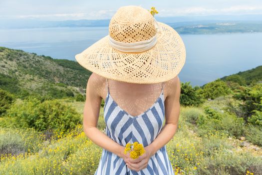 Young woman wearing striped summer dress and straw hat standing in super bloom of wildflowers, holding bouquet of yellow flowers in beautiful nature of Adriatic sea coastal nature of Croatia.