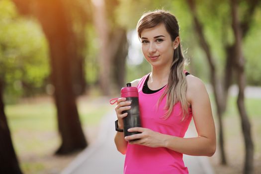 Beautiful fitness athlete runner woman drinking water in the park. Portrait face of a young woman holding a water bottle in summer. Forest