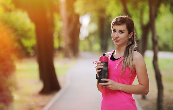 Beautiful fitness athlete runner woman drinking water in the park. Portrait face of a young woman holding a water bottle in summer. Forest