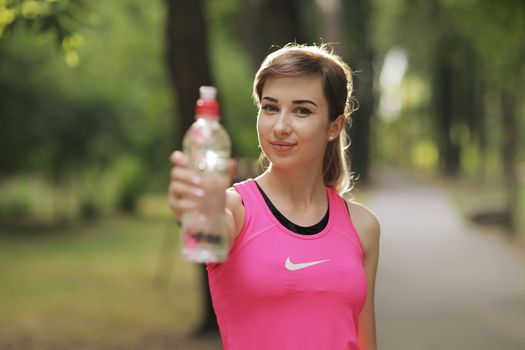 Beautiful fitness athlete runner woman drinking water in the park. Portrait face of a young woman holding a water bottle in summer. Forest