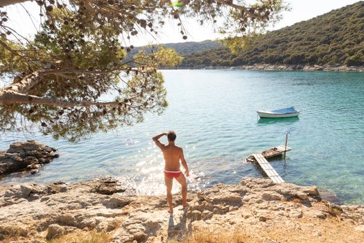 Rear view of man wearing red speedos tanning and realaxing on wild cove of Adriatic sea on a beach in shade of pine tree. Relaxed healthy lifestyle concept.