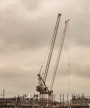 A construction crane and a building under construction made of concrete piles and iron, a construction site against a cloudy gray sky, vertical.