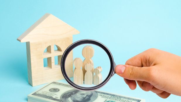 Magnifying glass is looking at the young family stands near a wooden house and with a bundle of money. The concept of a strong family, continuation. Loans for the purchase of affordable housing.